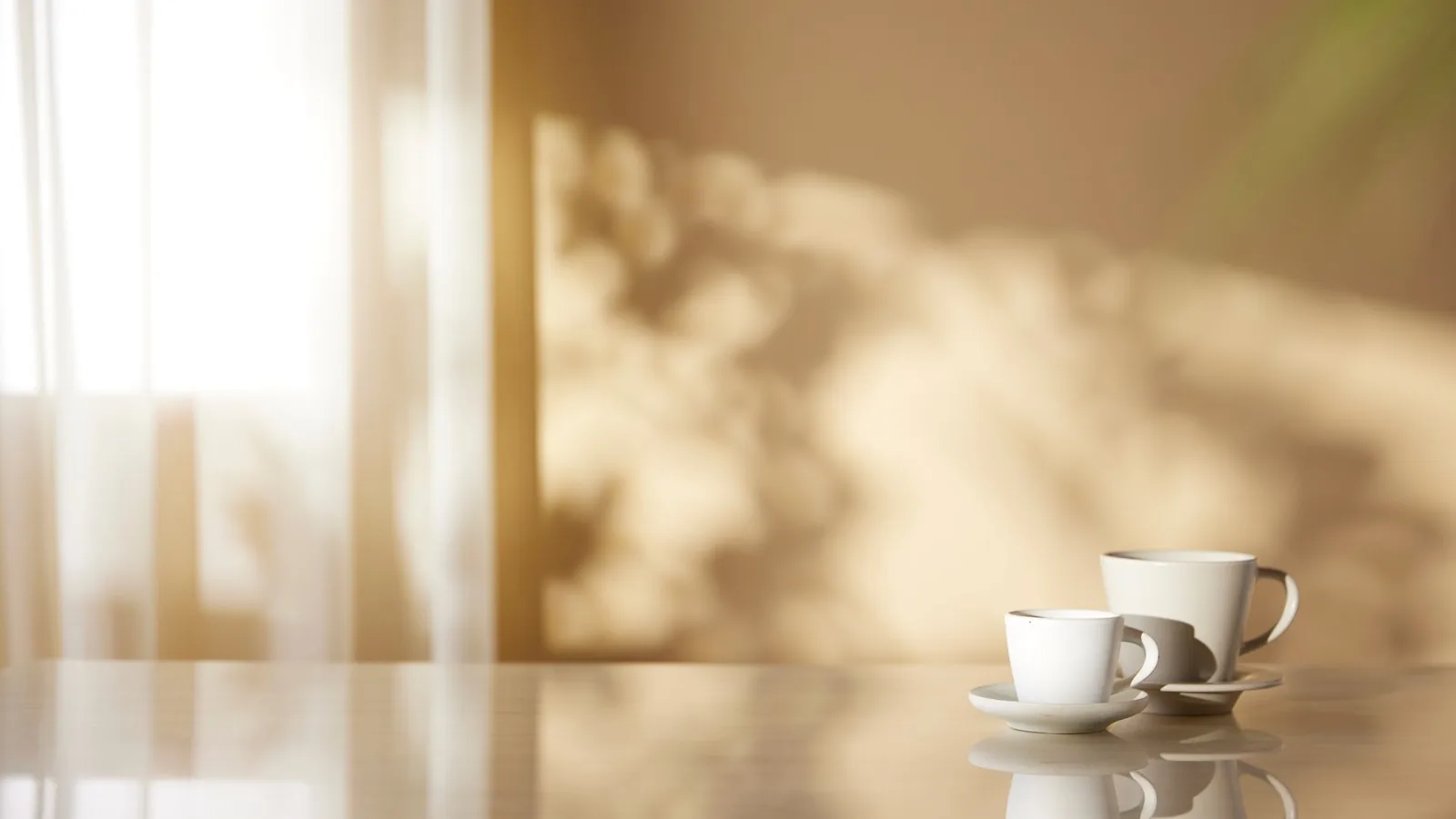 Two white cups on a polished table in a quiet room lit by soft morning light through a curtain