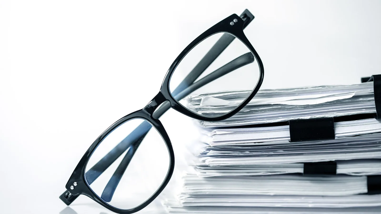 A pair of black reading glasses resting on a tall stack of clipped research papers
