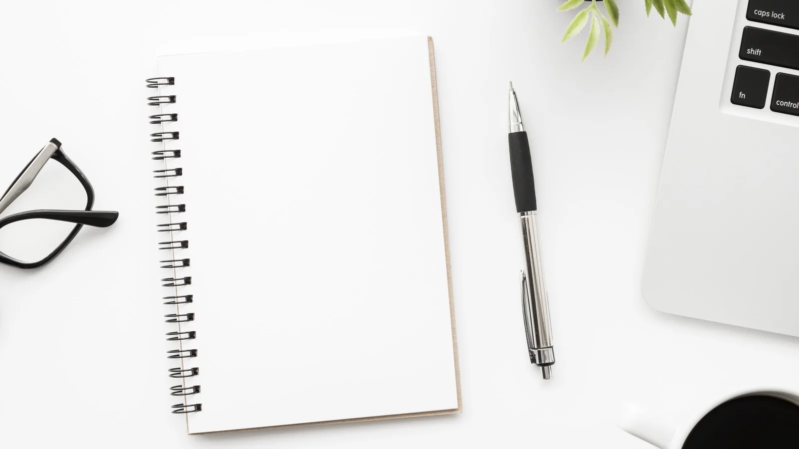 An open blank spiral notebook with a pen, reading glasses and a laptop on a white desk