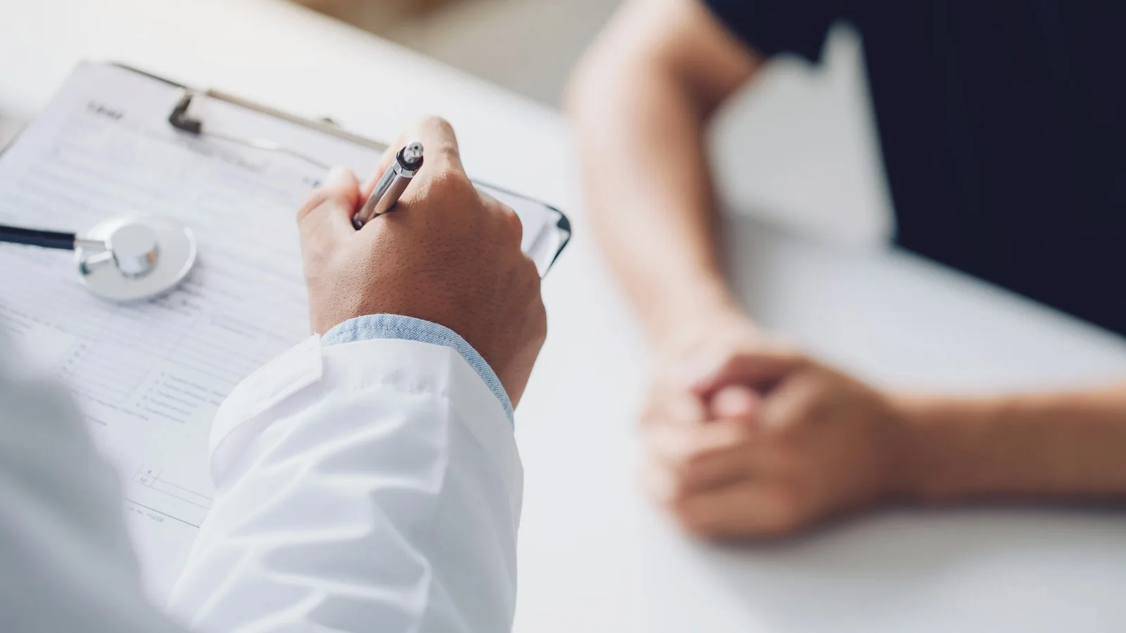 A clinician in a white coat writing on a clipboard across a desk from a patient's folded hands