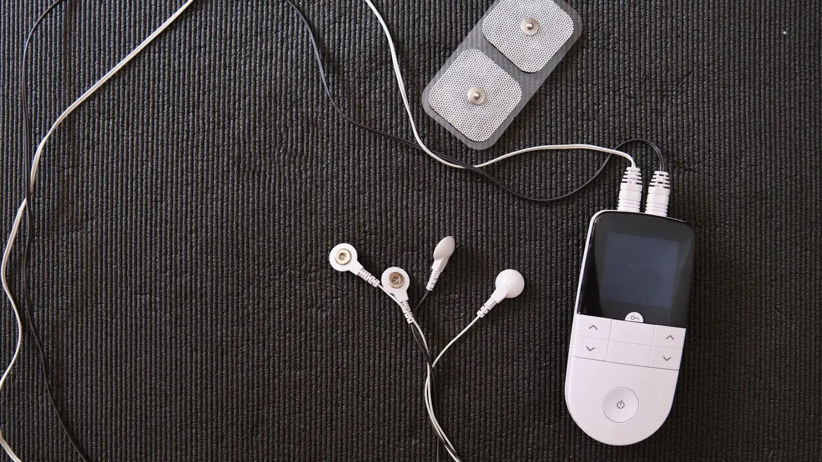 A small white TENS unit with its lead wires, ear clips and adhesive pads laid out on a dark mat